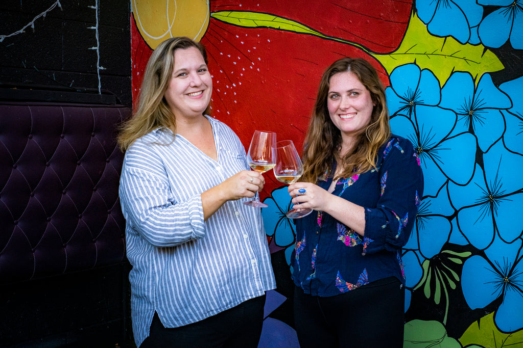 Julie and Allie standing in front of a brightly painted wall and holding wine glasses in a cheers position